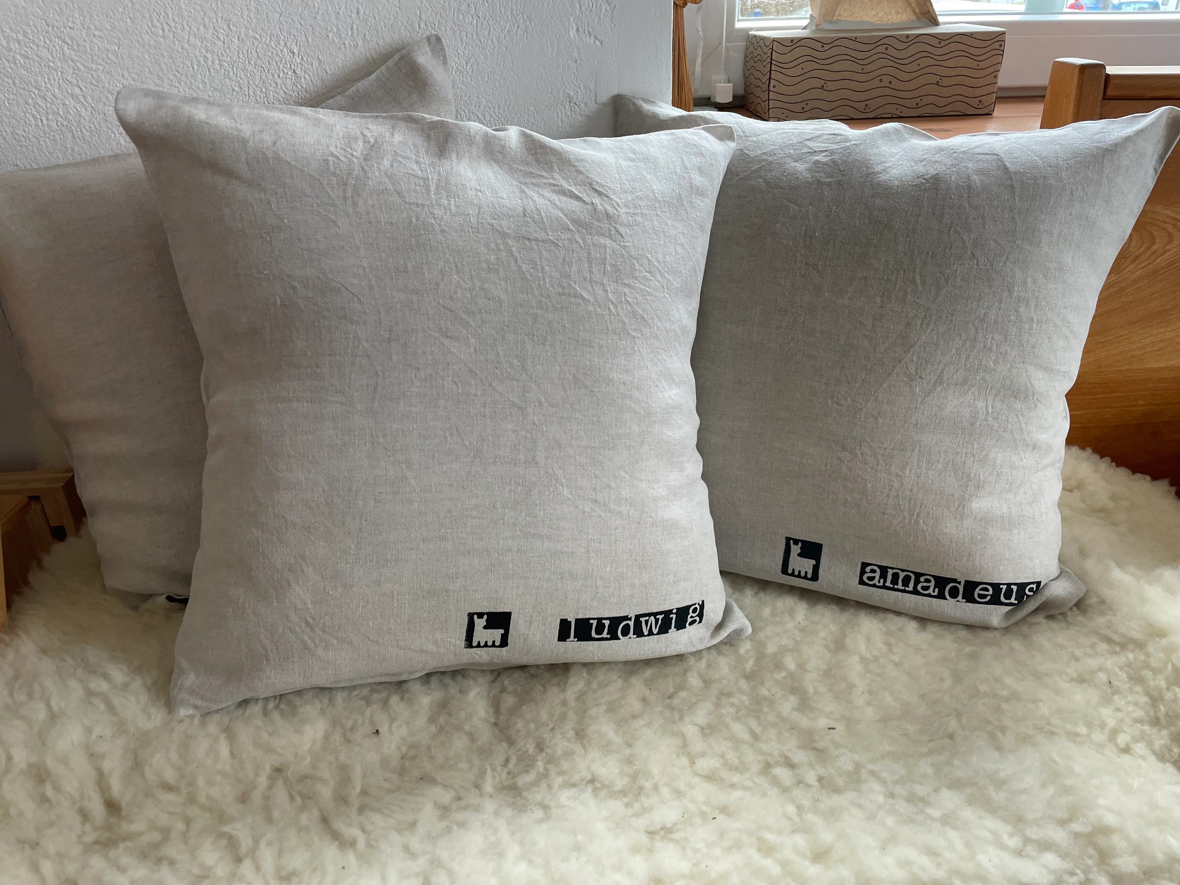Zwei Leinenpölster mit gestempeltem Alpaka-Logo und den Namen Ludwig und Amadeus, auf einem weißen Schaffell
