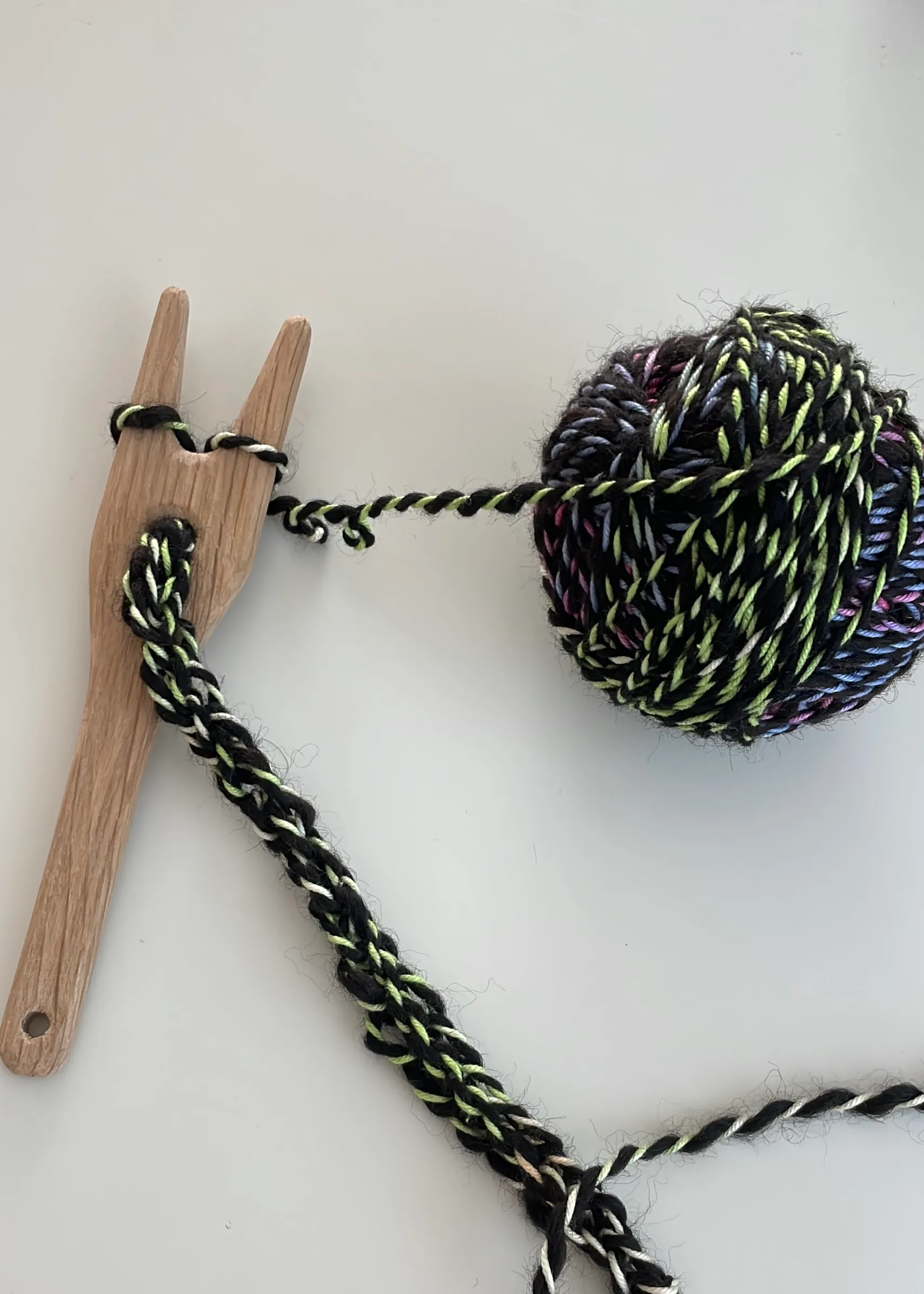Hölzerne Strickgabel mit einer angestrickten Kordel aus schwarzem Garn mit bunten Fäden, daneben ein Knäuel